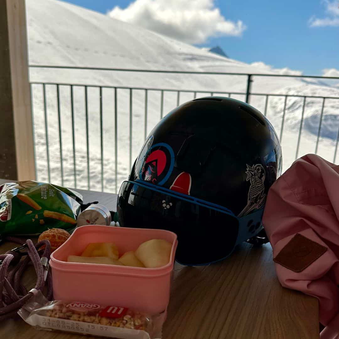 Ski helmet, jacket, snacks, and fruit laid out on a table at Roche de Mio, one of the most convenient La Plagne picnic spots with indoor warmth and snowy mountain views just outside.