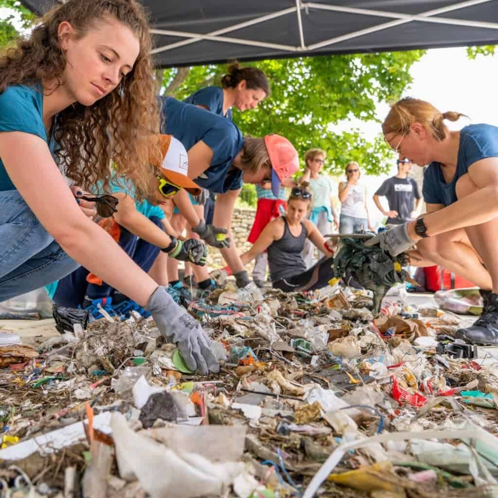 A group of volunteers sorts through a large pile of trash under a canopy during La Plagne clean up day, part of the Montagne Zéro Déchet initiative. They're wearing gloves and separating waste into categories with focus and teamwork.