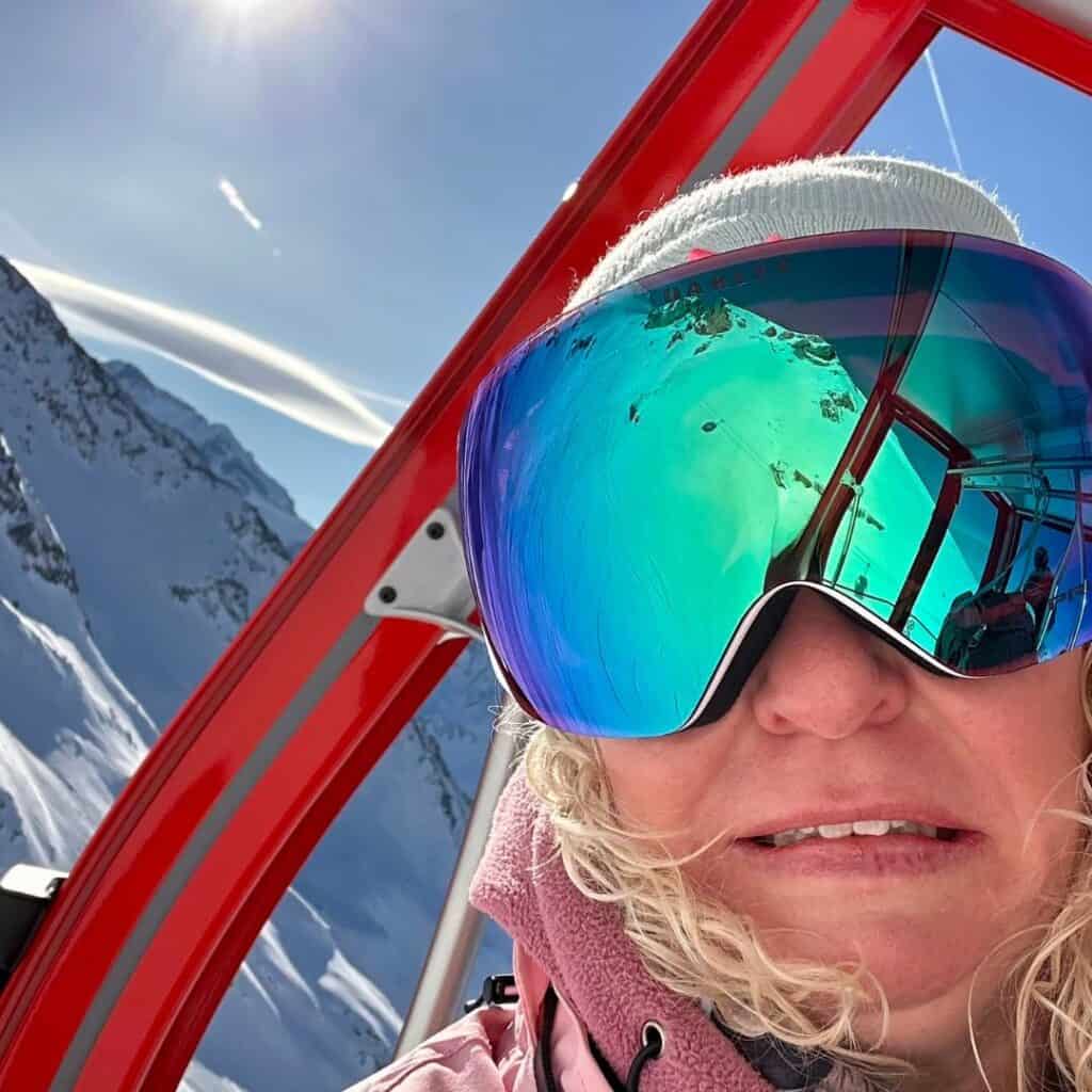 A woman with blonde hair and reflective ski goggles takes a selfie inside a red gondola, with snowy peaks and sunshine mirrored in her lenses. Her expression is a mix of excitement and nerves.