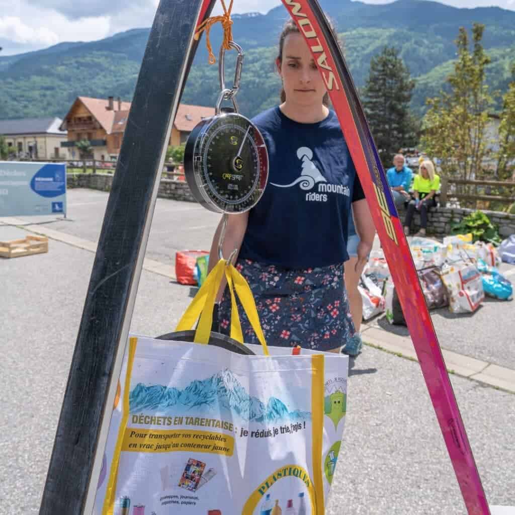 A volunteer from Montagne Zéro Déchet weighs a bag of recyclables using a hanging scale made from old skis during La Plagne clean up day, with the French Alps and collected bags in the background.