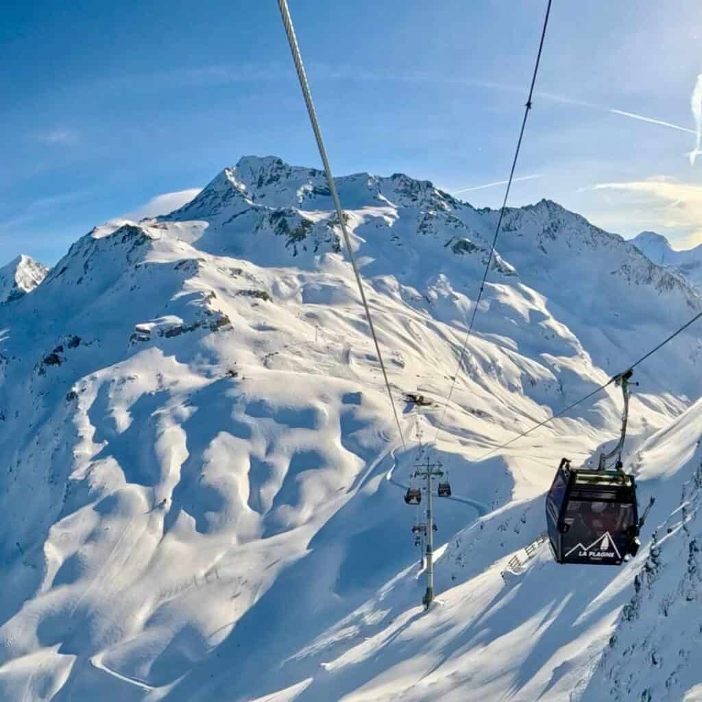 A black La Plagne gondola glides high above snow-covered mountains, suspended on a cable against a bright sky. The slopes below show winding ski runs and untouched powder.