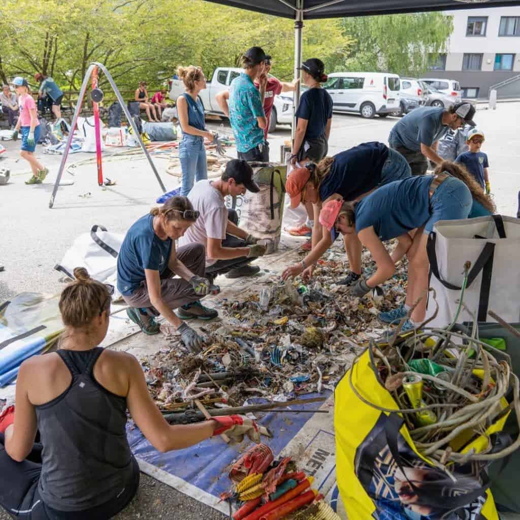 Teams of volunteers crouch under a tent carefully sorting litter into different bags during La Plagne clean up day, with a yellow bin of tangled wires and tools in the foreground.