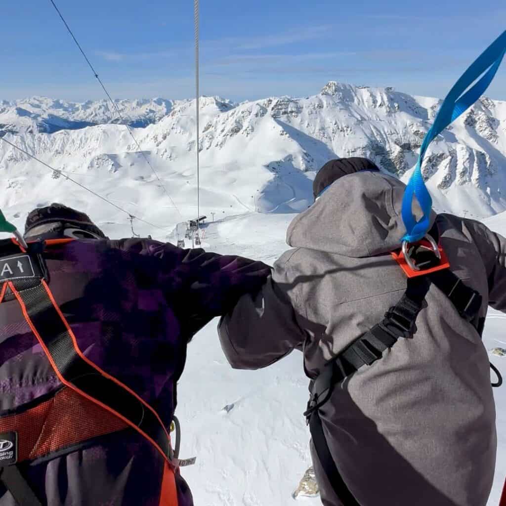 Two people wearing harnesses lean forward from a gondola, gazing over a dramatic snowy mountain landscape. Blue and green safety straps attach them to the red frame of the cable car.