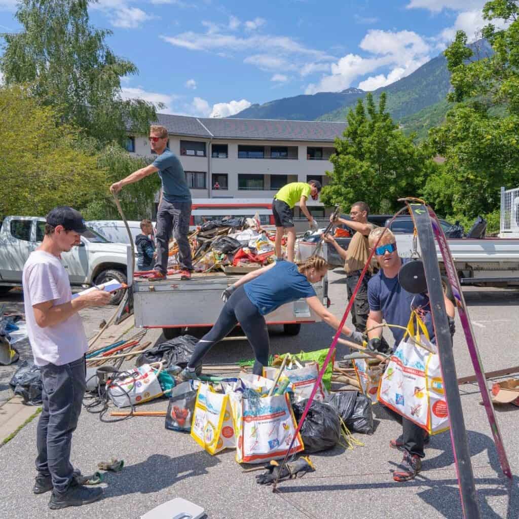 Volunteers unload and weigh collected waste on a sunny day during the Montagne Zéro Déchet event in La Plagne, with piles of sorted trash bags, colorful reusable bags, and a truck bed full of debris.