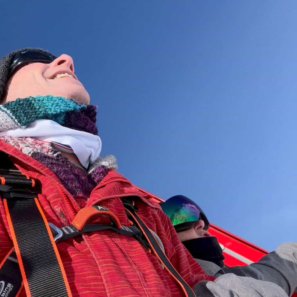 Close-up of two people looking up, bundled in scarves and snow gear with blue sky overhead. One wears a red jacket and a climbing harness, suggesting a high-altitude adventure.