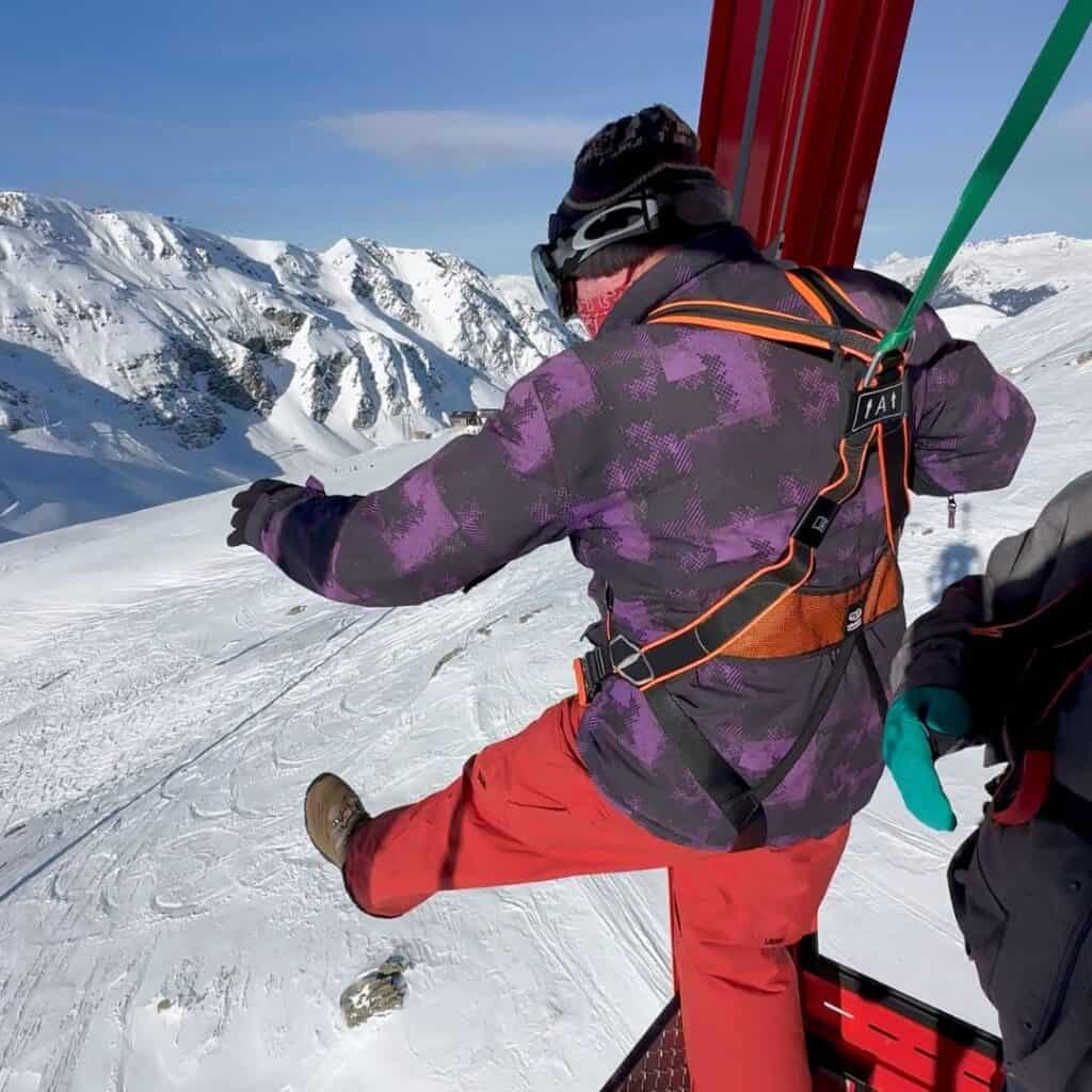 A person in a purple and black ski jacket and red pants steps out of a gondola, suspended by a harness over a snowy mountain, seemingly mid-action for a thrilling drop.