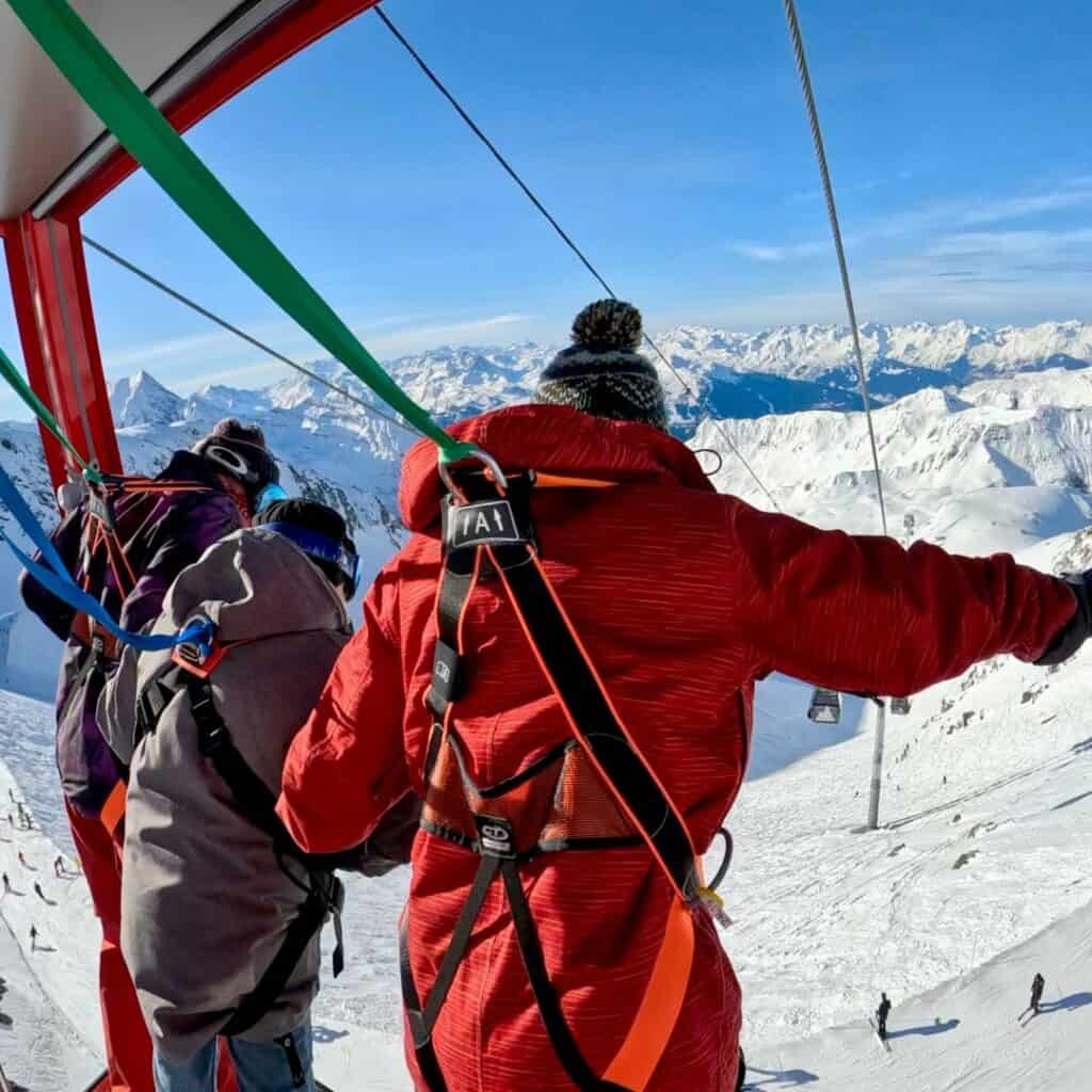 People in winter gear wearing harnesses are strapped into a red gondola, preparing for an adventurous descent over snowy alpine terrain. The expansive mountain range stretches into the distance under a clear blue sky.