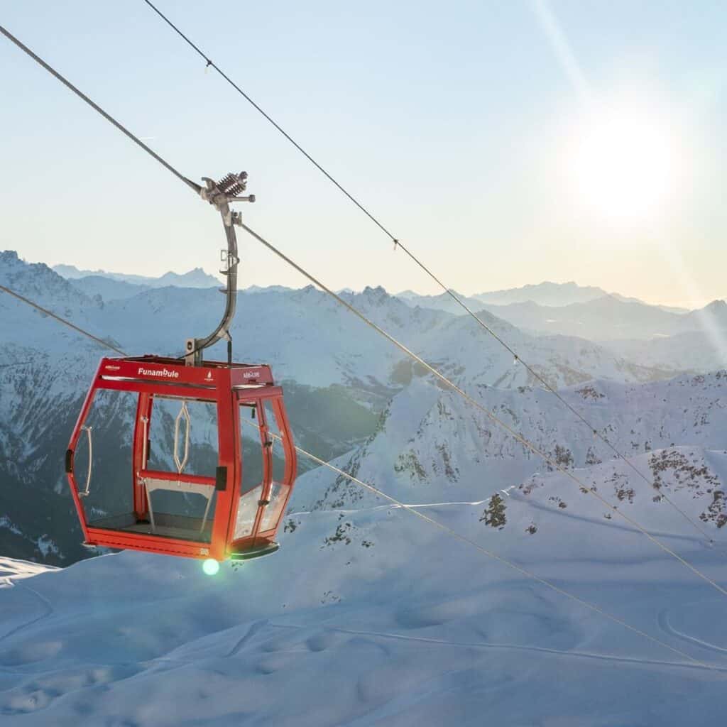 A bright red gondola carrying four people glides above a snow-covered mountain slope under a clear blue sky. Jagged rocky peaks peek out from the thick snow, adding a dramatic backdrop to the alpine scene.