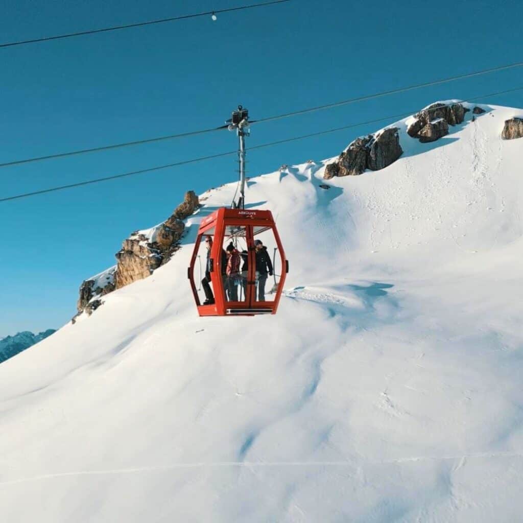 A red gondola lift suspended over a pristine snowy mountain slope, carrying skiers up the mountain at La Plagne under a clear blue sky.