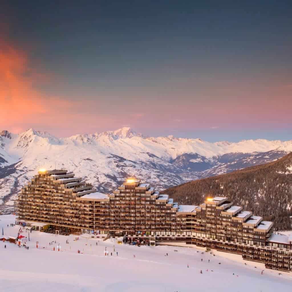 Iconic tiered architecture of Aime 2000 at sunset, glowing under soft lights with snow-covered mountains and pink twilight skies in the distance.