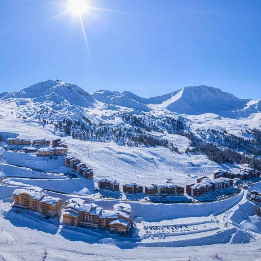 Aerial view of snow-covered slopes and clustered lodges under bright sunshine in Plagne Villages, one of the 11 La Plagne villages, with panoramic mountain peaks in the background.