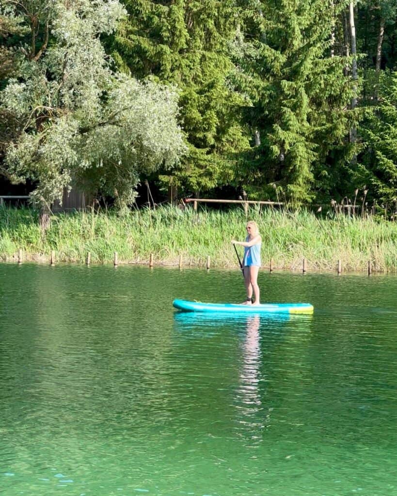 A woman stands on a bright blue and yellow Fanatic Fly Air paddle board, gliding across a calm green lake near La Plagne’s Plan d’Eau. She’s paddling in front of a lush forest shoreline on a sunny summer day, enjoying the peaceful water.