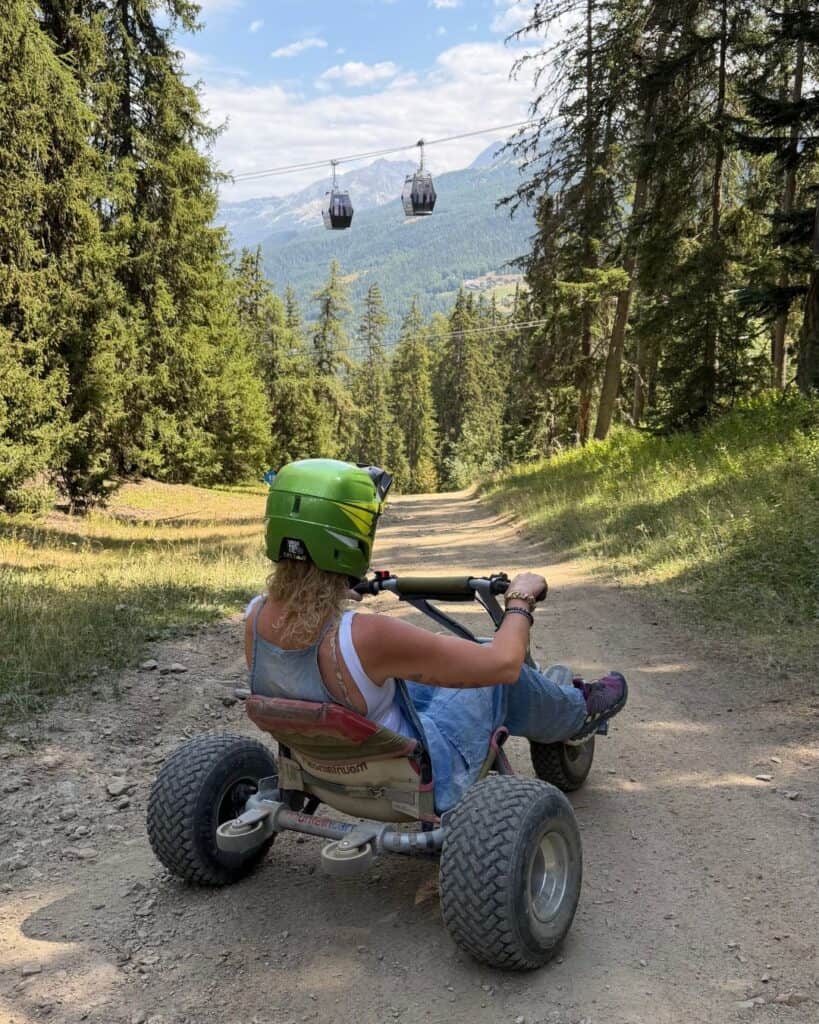 Mountain carting down a forest track in Plagne Centre, La Plagne, with gondola cabins and mountain views in the background.