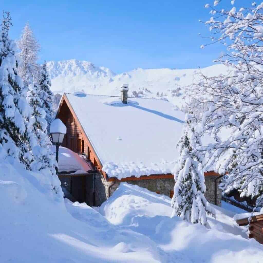 Snowy Will I Ski Chalet in La Plagne with Mountain View – A charming chalet with a snow-covered roof nestled among snowy trees, with breathtaking alpine mountains in the background under a clear blue sky.