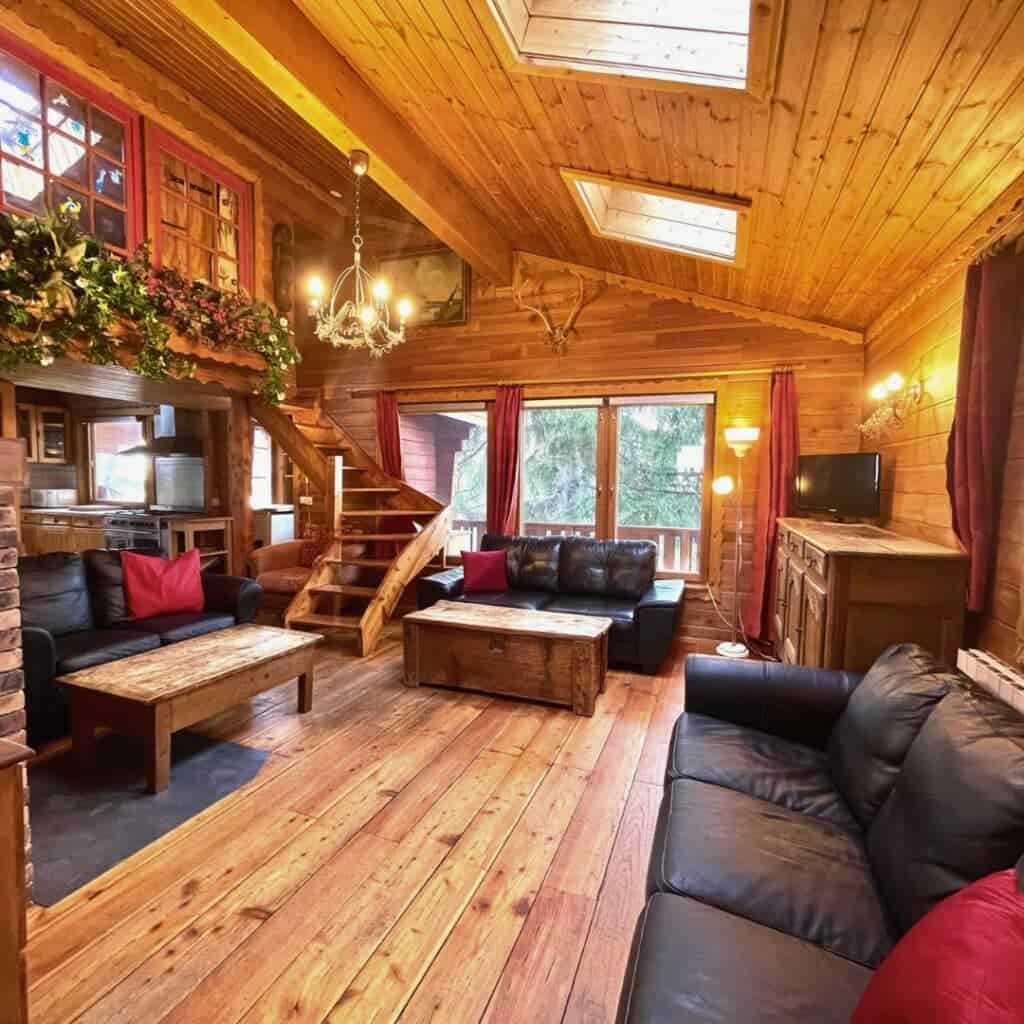 Cosy Will I Ski La Plagne Chalet Living Room – "A warm, wooden chalet living room with black leather sofas, red accent pillows, a rustic coffee table, and a chandelier. Large windows and skylights fill the space with natural light.