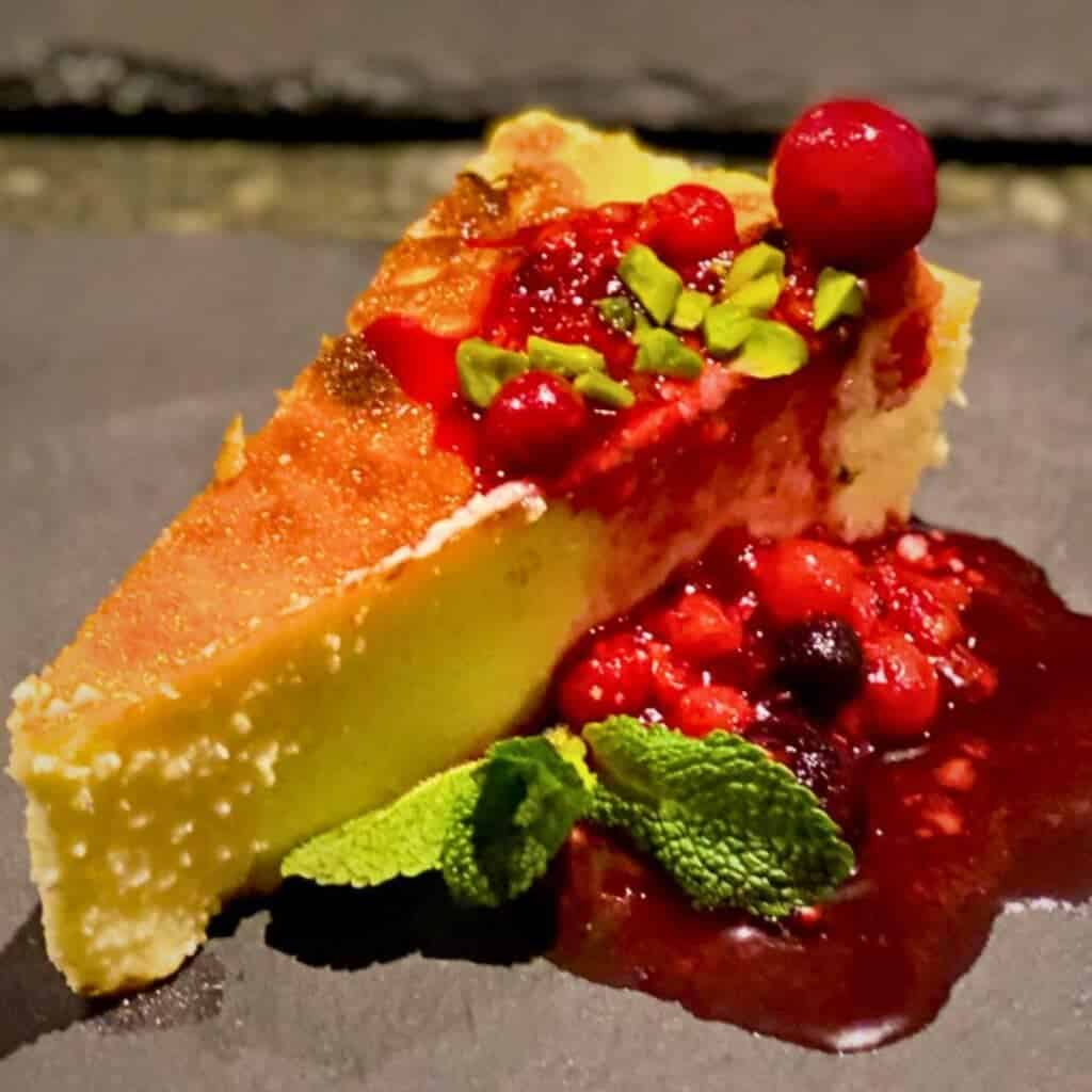 A slice of creamy cheesecake with a golden-brown top, garnished with a vibrant berry compote and crushed pistachios. The dessert is plated on a dark surface, with fresh mint leaves adding a touch of green to the rich red sauce. Credit: Alpoholics Ski Chalets La Plagne