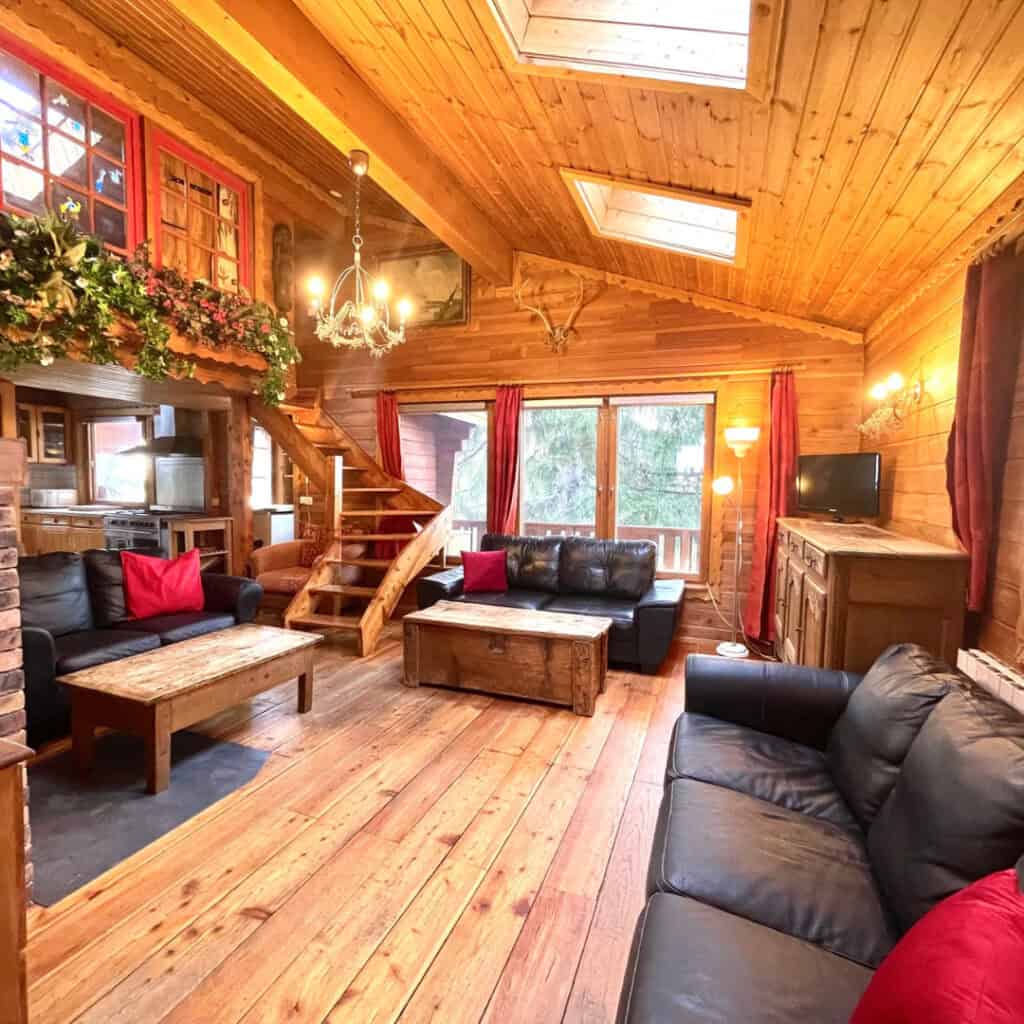 Chalet Caché Living Room – Cosy alpine living room with wooden interiors, black leather sofas, a rustic wooden coffee table, and warm lighting. A staircase leads to an upper level, while large windows and skylights let in natural light.