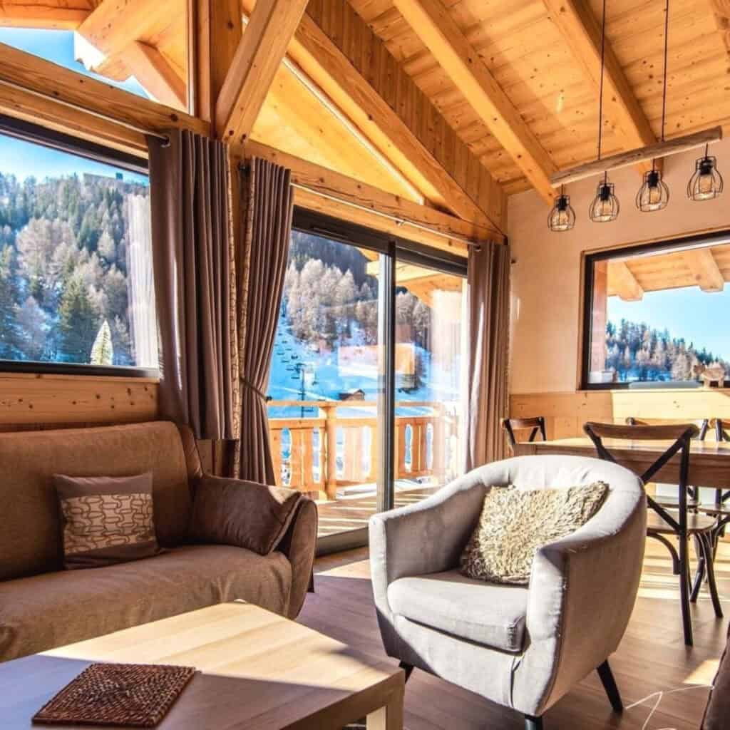 (Chalet Choucas Interior with Mountain View) – Bright and modern chalet interior featuring a plush armchair, a cozy sofa, and a wooden dining table. Large windows open to a balcony with stunning views of the snowy slopes and pine trees.