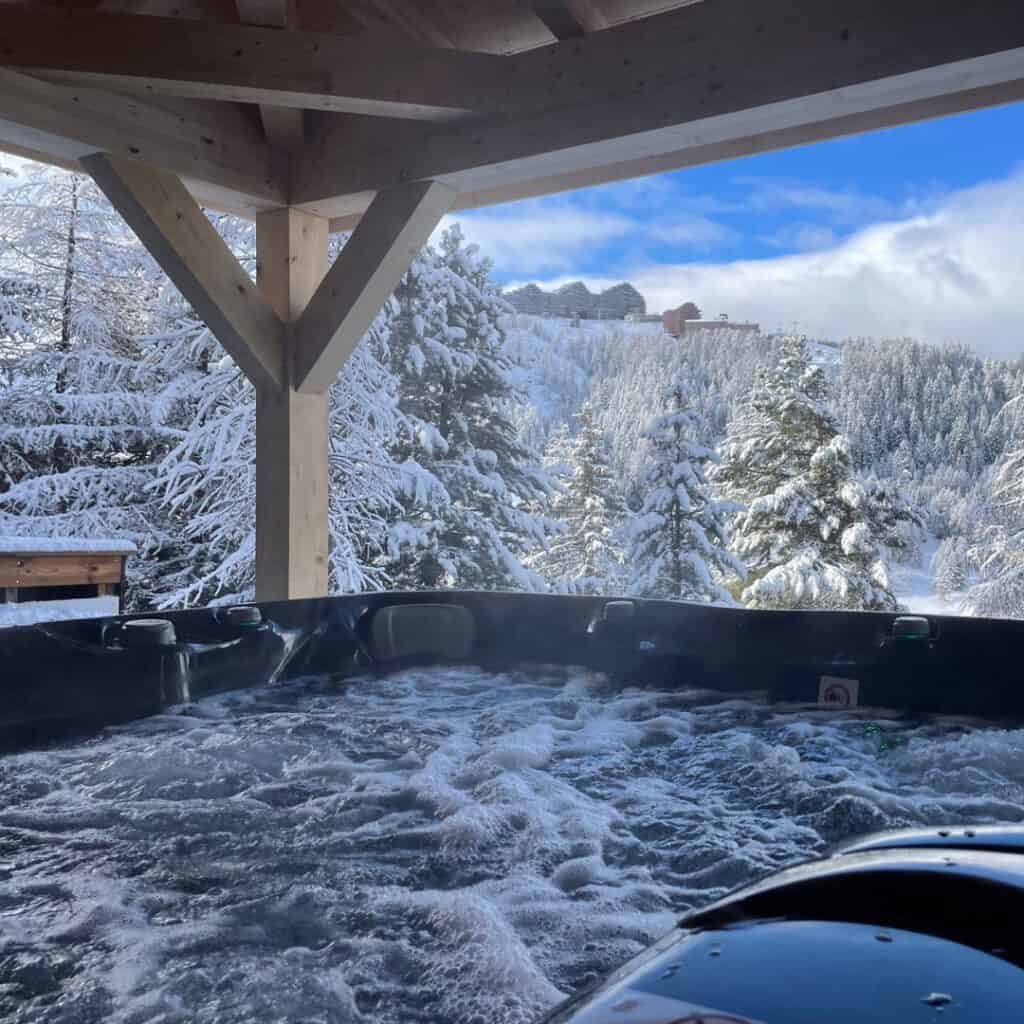 (Chalet Genepy Hot Tub with Snowy Mountain View) – Steaming outdoor hot tub under a wooden pergola, overlooking a breathtaking winter landscape of snow-covered trees and mountains.