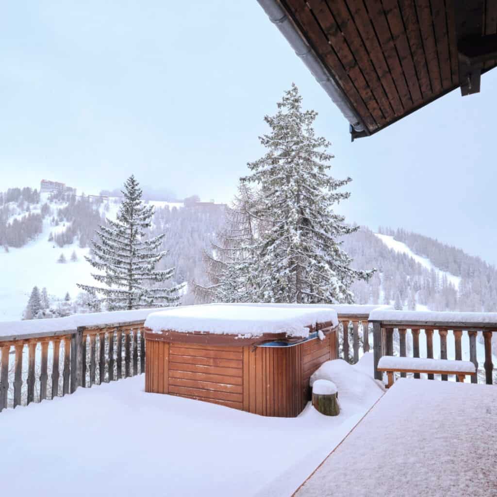 (Chalet Oursons Snowy Hot Tub Deck) – A wooden hot tub covered in fresh snow sits on a chalet’s deck, surrounded by a winter wonderland of snow-covered pine trees and ski slopes.