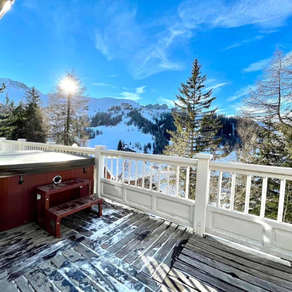 (Chalet Plume Deck with Mountain View) – A chalet deck with a steaming hot tub and a wooden floor dusted with snow, overlooking a breathtaking alpine landscape with bright blue skies and sunlit peaks.