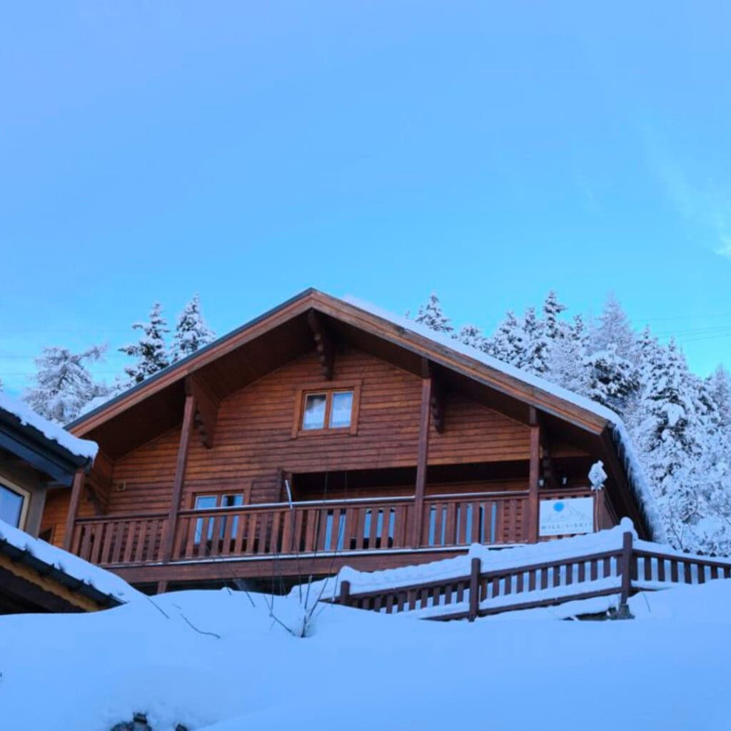(Chalet Zibeline Exterior in the Snow) – Traditional wooden chalet with a snow-covered roof and balcony, nestled among frosty pine trees under a clear blue sky.