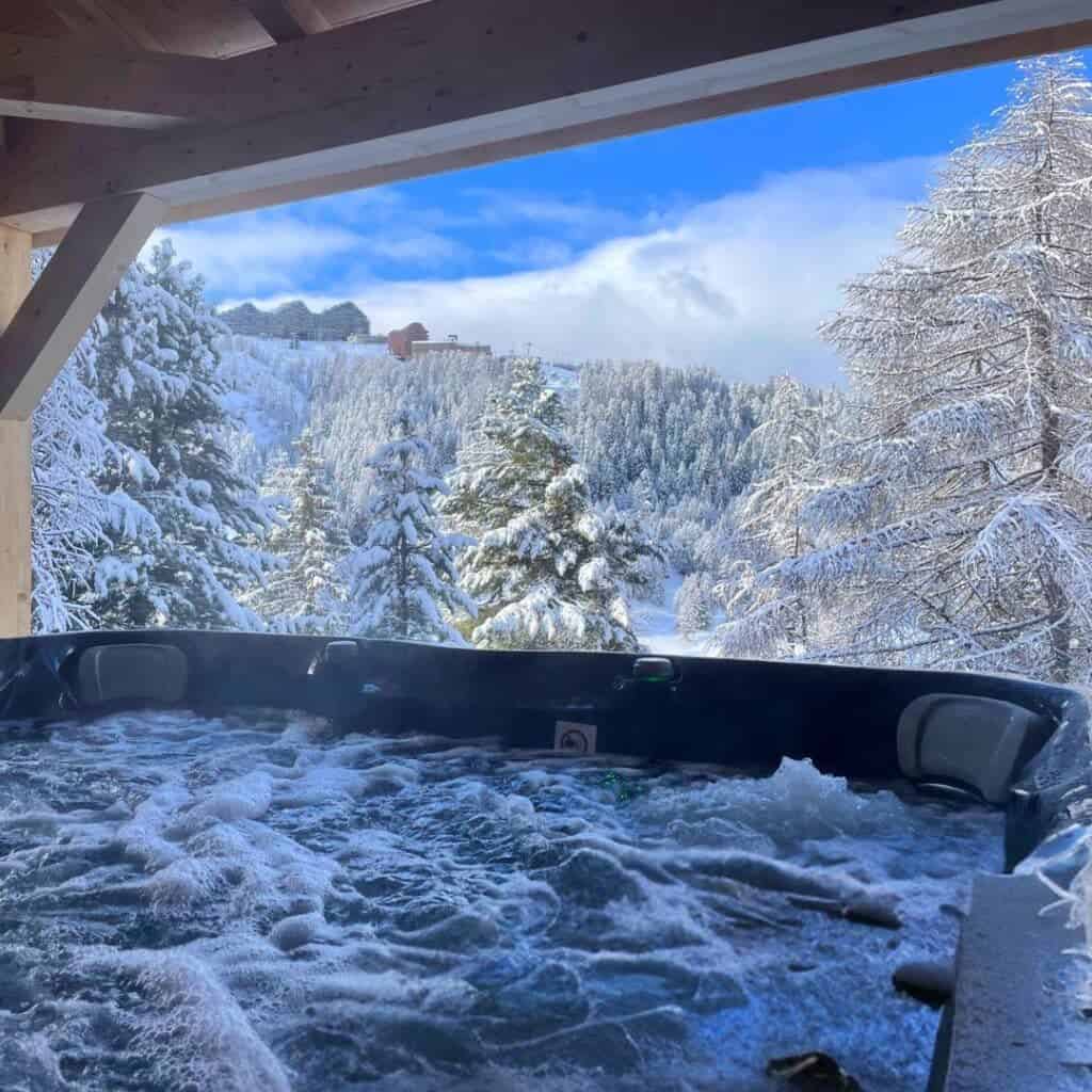 Will I Ski La Plagne Chalet Hot Tub with Snowy Mountain View – An inviting outdoor hot tub with steaming water, set against a stunning winter landscape of snow-covered trees and mountains beneath a bright blue sky.