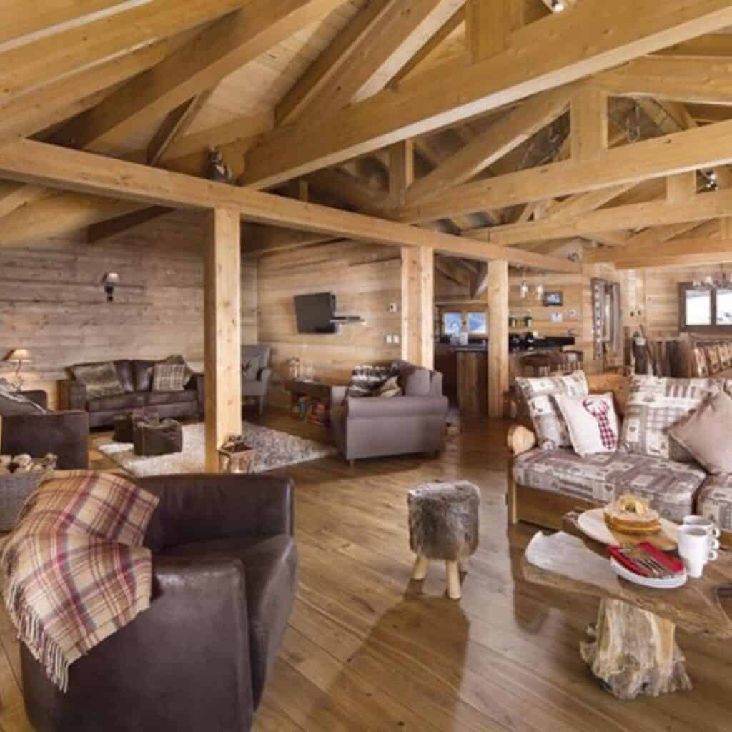 A cozy, rustic chalet-style living room with exposed wooden beams, wooden walls, and a high ceiling. The space is furnished with plush sofas, armchairs, and a mix of modern and traditional decor, including plaid blankets and patterned cushions. A wooden coffee table with a tree-stump base holds a breakfast setting with coffee and pastries, adding to the warm, inviting atmosphere.