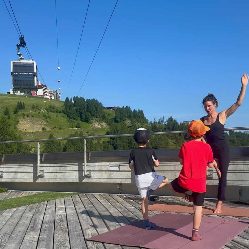 Instructor and two children balancing in tree pose during an outdoor yoga session, with a large gondola passing overhead and lush green hills in the background.