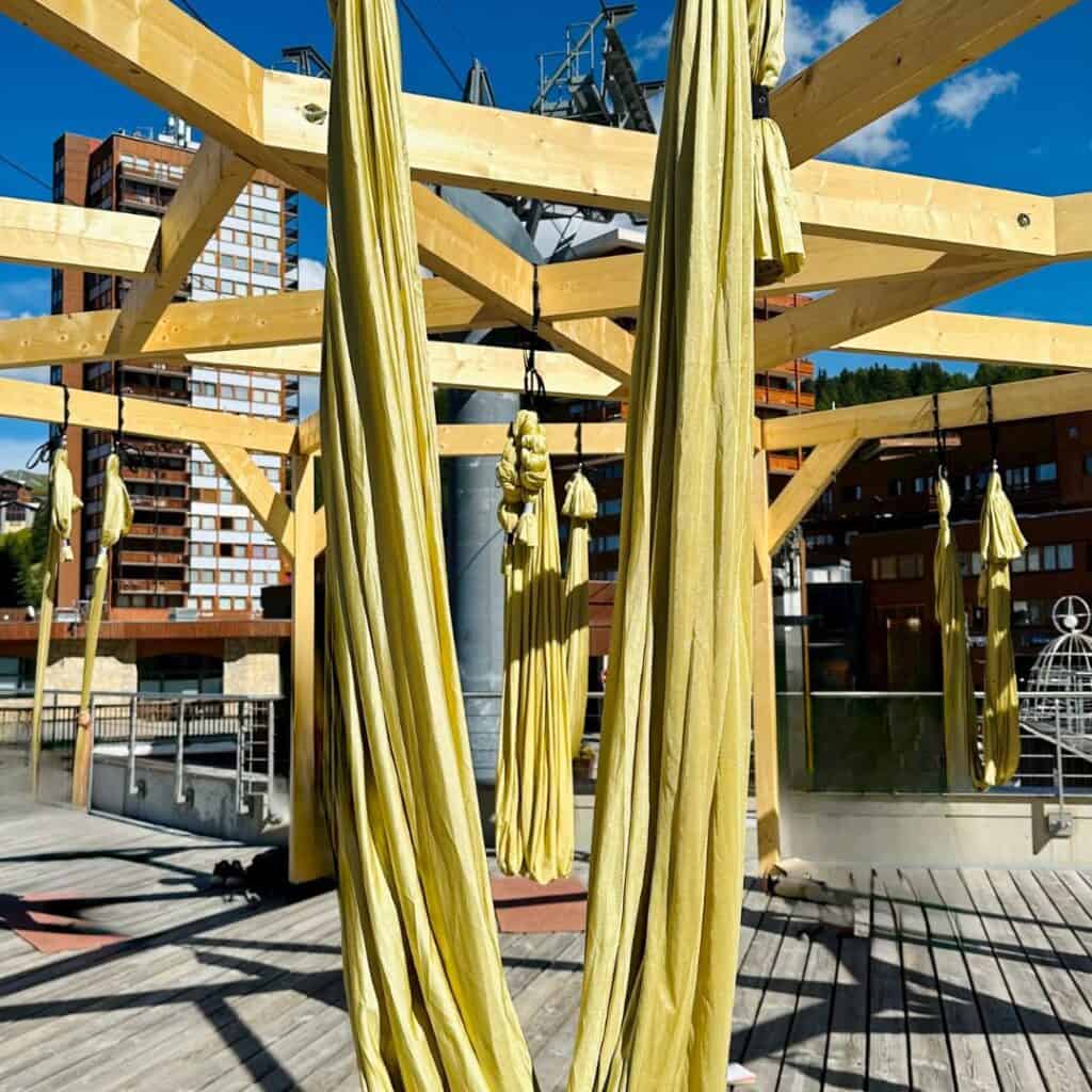 Group of yellow aerial yoga hammocks hanging from a sturdy wooden frame, set against a modern alpine resort backdrop with cable infrastructure and blue skies.