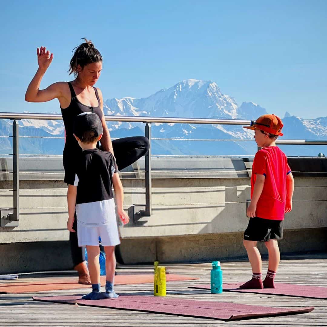 Yoga instructor demonstrating a tree pose in front of two children standing on mats, with water bottles at their feet and majestic snow-covered mountains in the distance.