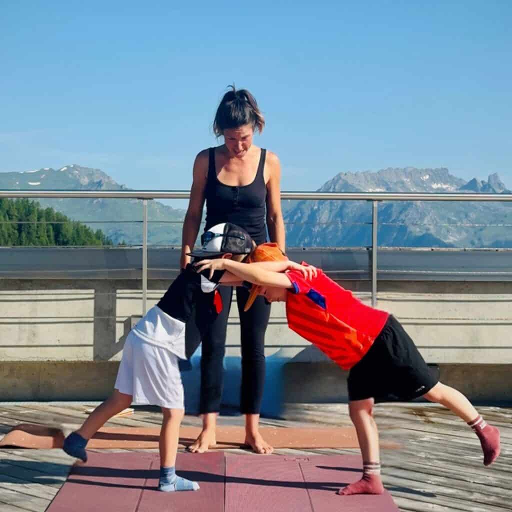 Two boys performing a leaning partner stretch while standing on yoga mats, assisted by an instructor, with green mountains and clear blue skies behind them.