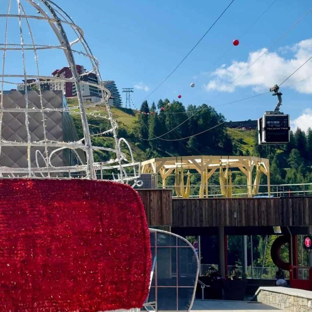 View of a gondola lift at La Plagne passing above a terrace with yellow aerial yoga hammocks, with festive decorations and grassy ski slopes in the background.