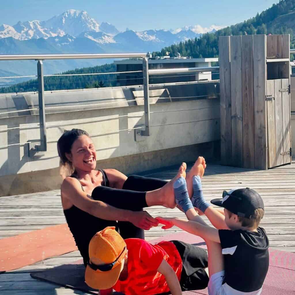 Smiling woman and two boys enjoying a seated partner yoga pose on a mat, interlocking feet and hands, surrounded by wooden decking and mountain views.