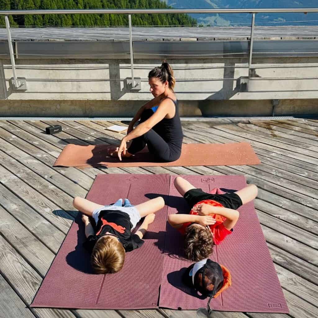 Two children lying on yoga mats in a resting position while an instructor sits in meditation on her mat nearby, with forested slopes and a panoramic view behind them.