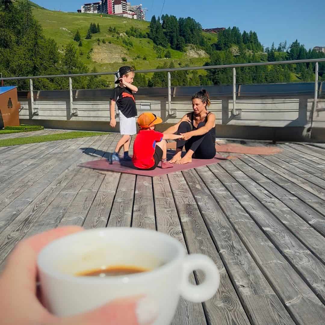 A woman and two kids engaging in a yoga session on a wooden deck with a grassy hill and alpine buildings in the background, while a hand holding a cup of espresso is visible in the foreground.