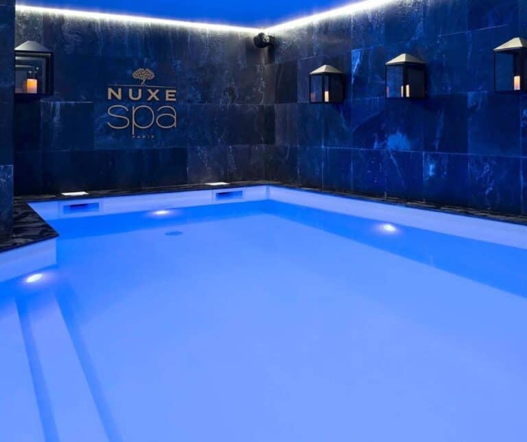 A luxurious indoor pool at NUXE Spa with dark slate tile walls, soft blue lighting, and candle-lit lanterns mounted on the walls.