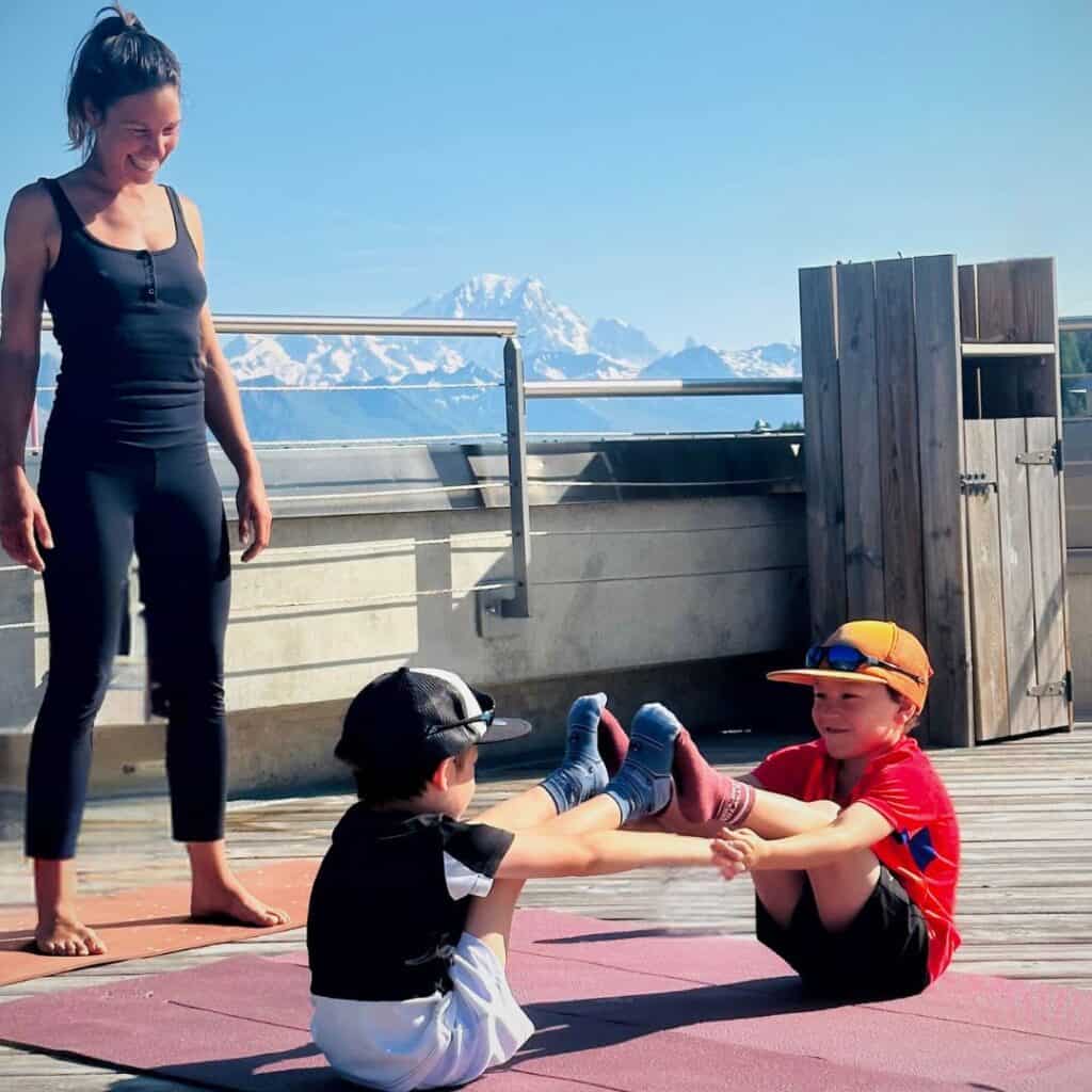 Two children practicing a partner yoga pose on a mat, holding each other’s feet while balancing, with an instructor smiling beside them and snow-capped mountains in the background.