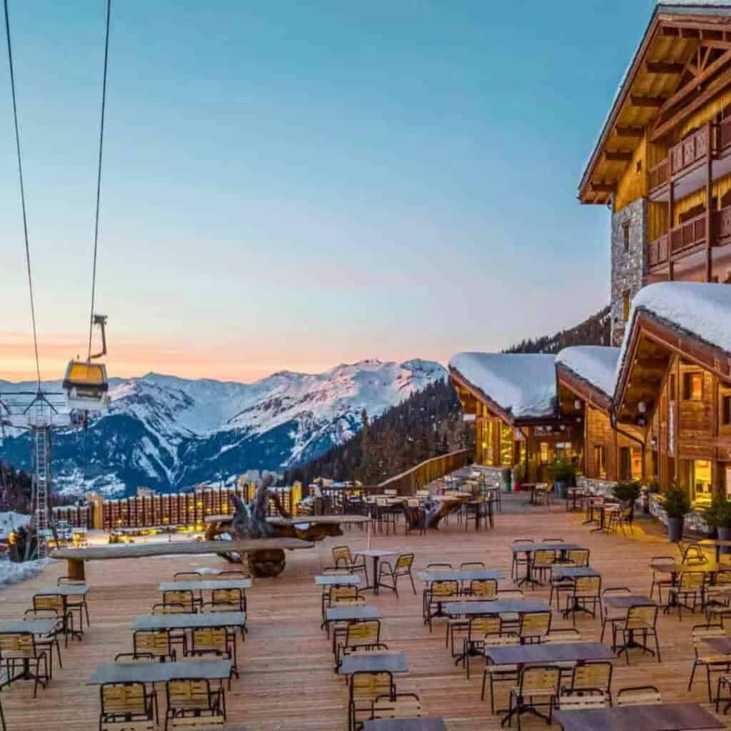 Hotel Carlina Restaurant Terrace La Plagne: A stunning outdoor terrace at a ski resort, overlooking a snow-covered mountain range during sunset. The wooden deck is lined with tables and chairs, while cozy chalet buildings with snow-covered roofs and glowing windows create a warm atmosphere. A ski lift in the foreground ascends into the distance, completing the picturesque alpine setting.