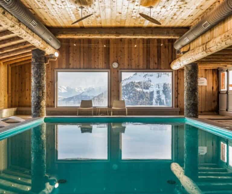 Cozy indoor pool framed by warm wooden walls and beams, with large windows showcasing a snowy alpine mountain view and lounge chairs in front.