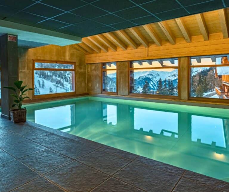 A warm-lit indoor pool with rustic wooden beams and wide windows showcasing a panoramic snowy mountain view.