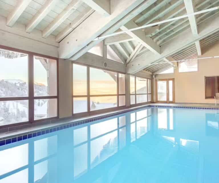 A serene indoor pool with a light blue ceiling and exposed beams, surrounded by large windows framing a pastel sunset over snow-covered rooftops and mountains.