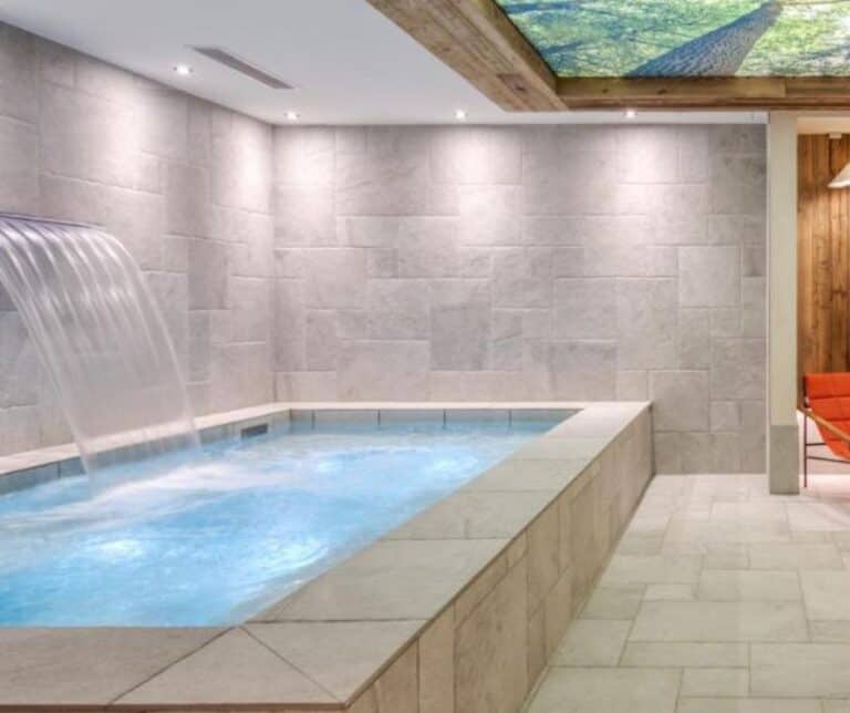 A minimalist spa pool with a stone-tiled floor and a wide water fountain spilling from the wall into a bubbling rectangular pool, next to orange lounge chairs and wooden accents.