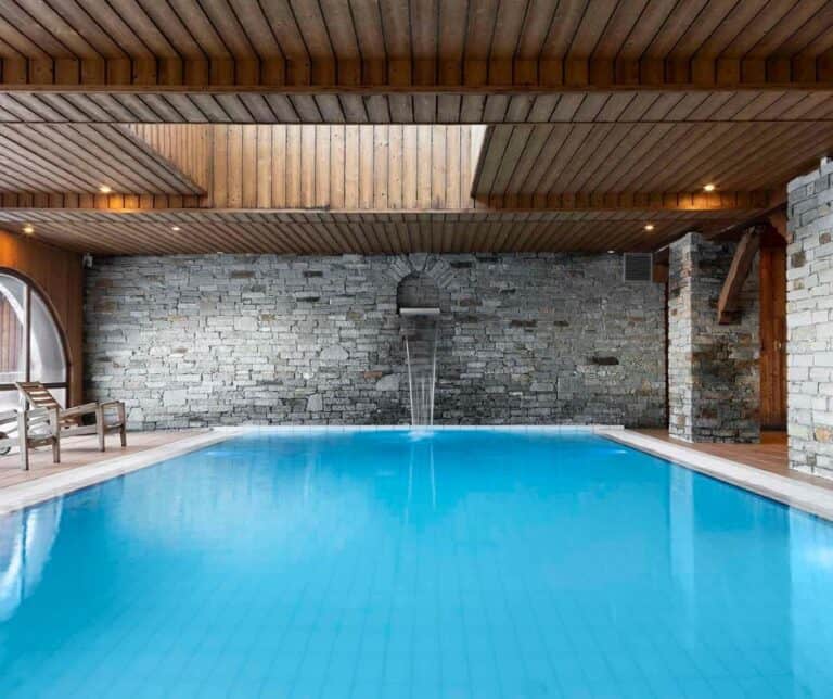 Indoor swimming pool with rustic stone walls and wood-paneled ceiling, featuring a gentle water spout and wooden deck chairs on the side.