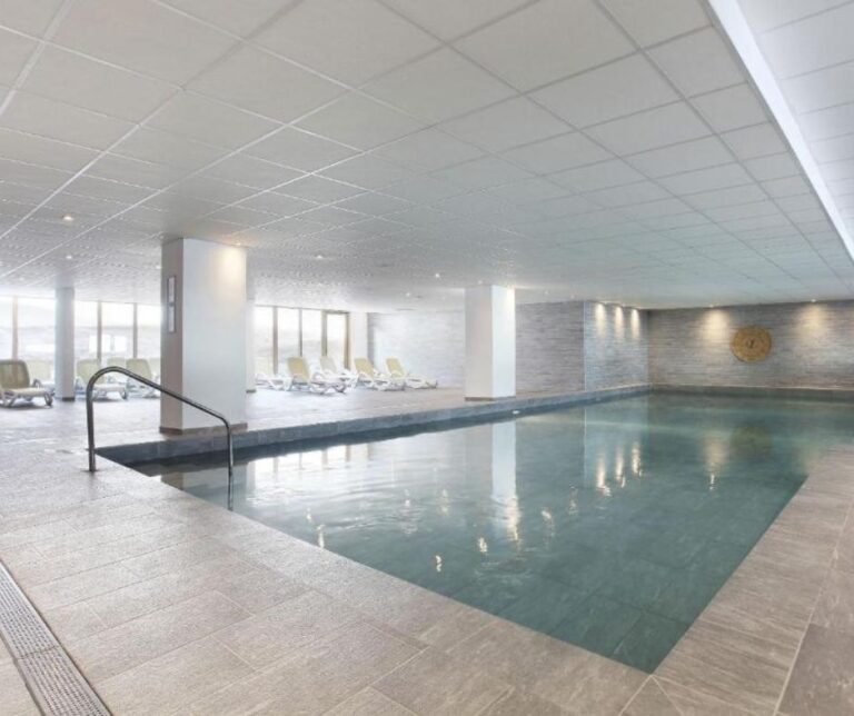 A modern indoor pool area with soft lighting, grey tiled walls and floors, and multiple lounge chairs near large windows.