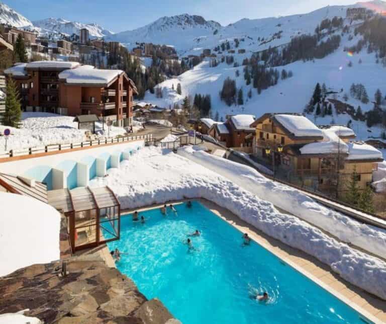 An outdoor heated pool nestled in a snowy mountain resort village with swimmers enjoying the warm water amidst snow-covered rooftops and ski slopes.