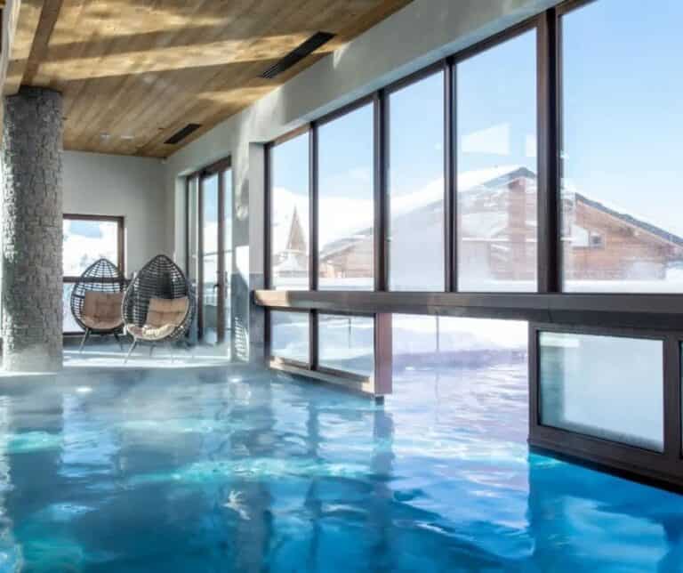 An elegant indoor pool with large floor-to-ceiling windows offering snowy mountain views; two cozy egg-shaped wicker chairs with cushions sit in the corner beside a stone column.