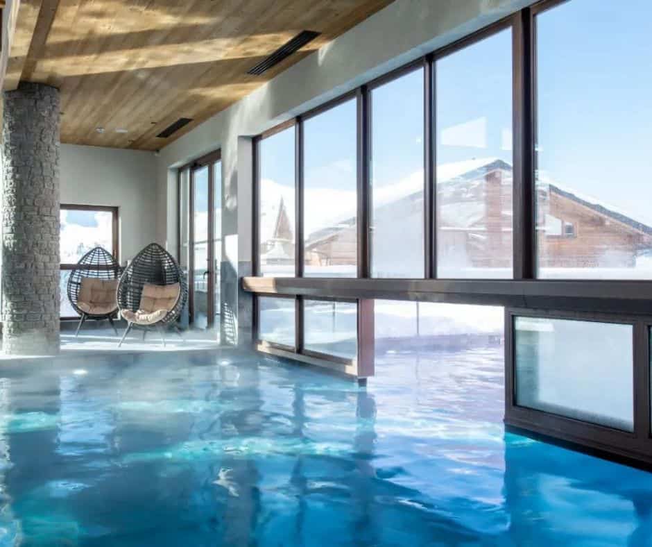 An elegant indoor pool with large floor-to-ceiling windows offering snowy mountain views; two cozy egg-shaped wicker chairs with cushions sit in the corner beside a stone column.