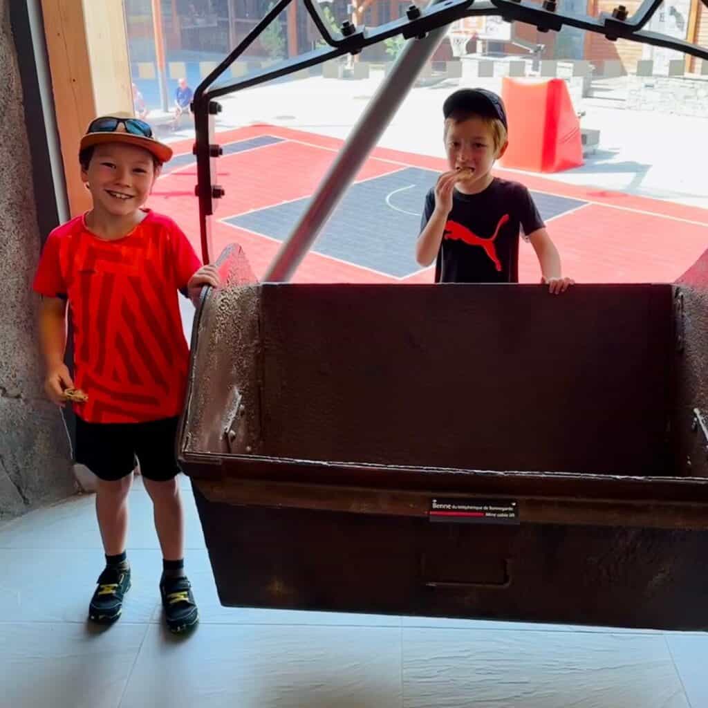 Two young boys standing by a large mining container inside a museum, with a basketball court visible outside through the window.