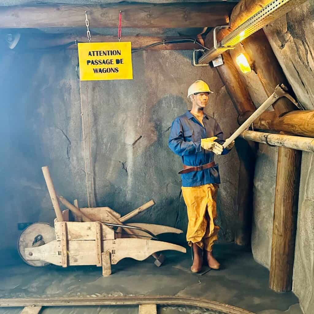 Life-size mannequin in a hard hat and miner’s uniform holding a tool inside a mining exhibit, next to a wooden cart and a yellow sign reading "Attention Passage de Wagons."